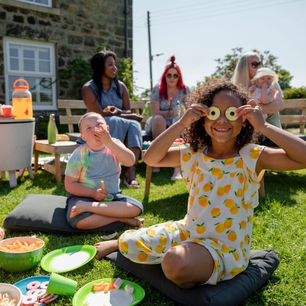 A sunny picnic scene with a boy eating and a girl in summer dress holding partyring biscuits over her eyes and smiling