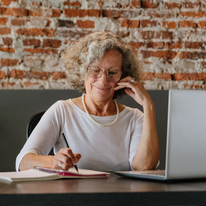 An older woman writes down notes as she works at her laptop