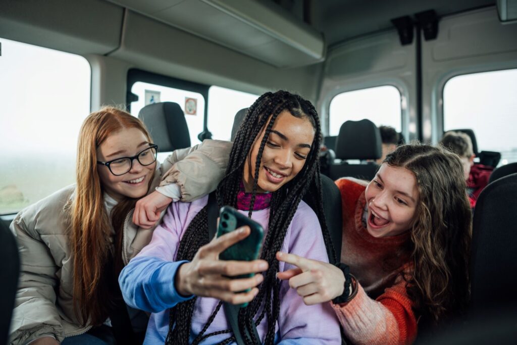 A group of teenage girls on a minibus laugh and point to the smartphone that the girl in the middle is holding.