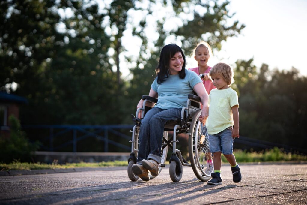 Mum in a wheelchair outdoors with a young girl pushing her wheelchair and younger boy walking beside her