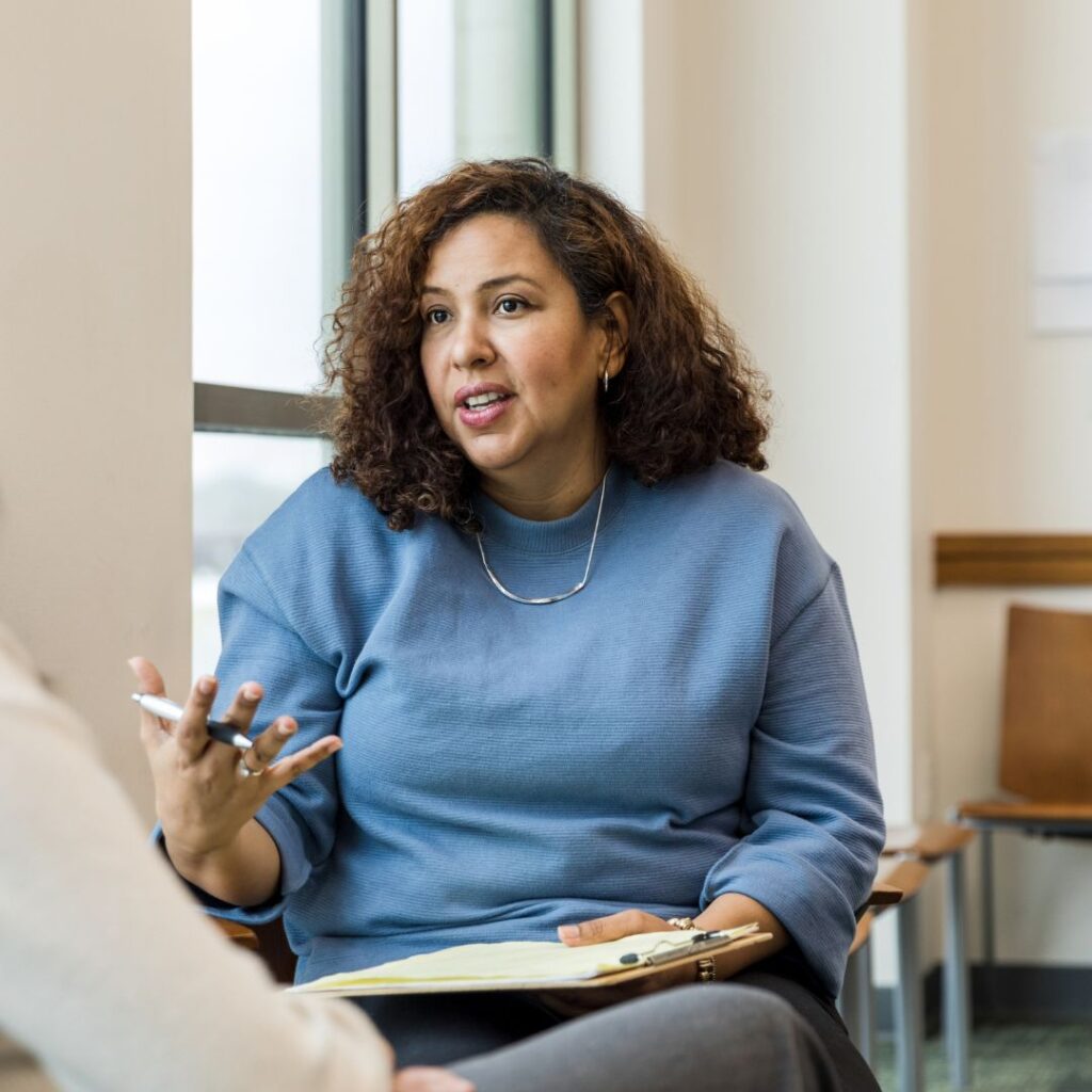 A professional woman with a clip board and pen talks to another woman sitting just out of sight