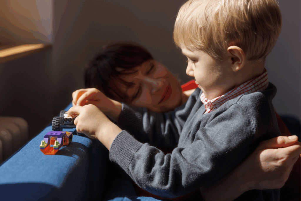 A woman with brown hair gazes adoringly at her young blonde son, who is sat in the sunlight playing with a lego car.