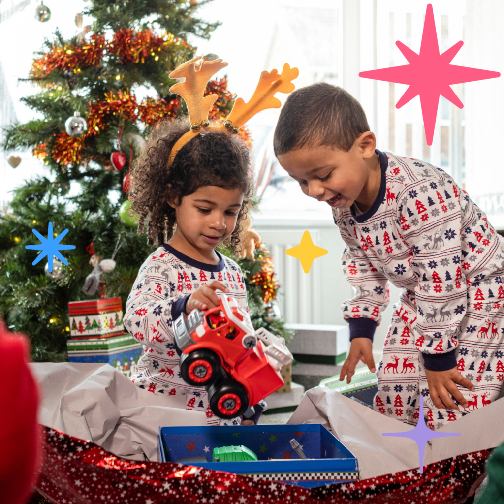 Two young children in Christmas pyjamas sit in front of a Christmas tree opening a present of a red tractor.